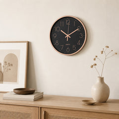Modern black wall clock with rose gold details displayed above a wooden sideboard